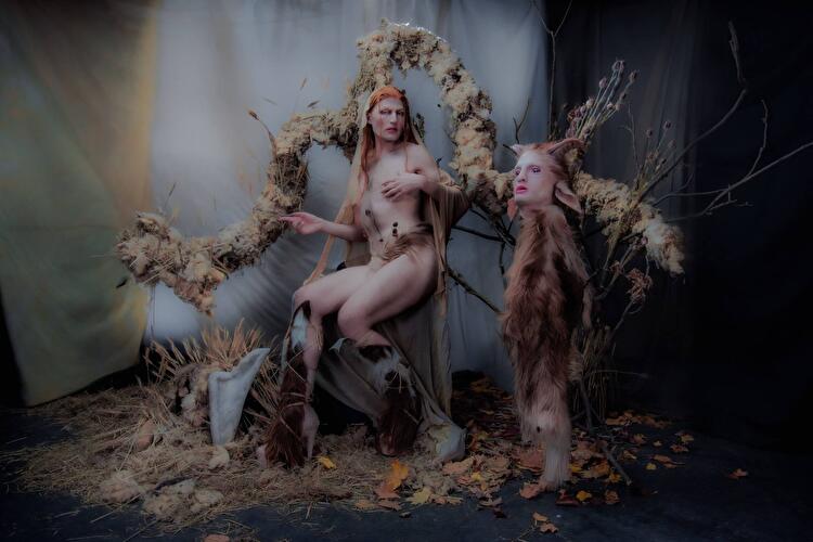 Inszenierte Fantasiefigur in naturverbundener Kulisse mit Fell, Zweigen und Blüten, Teil einer künstlerischen Performance.