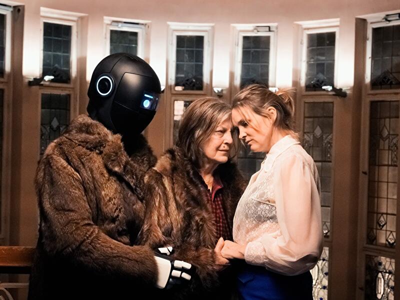 Zwei Personen und eine humanoide Roboterfigur in Mänteln stehen in einem beleuchteten Innenraum zusammen.