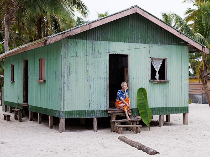 Person sitzt vor einem einfachen Stelzenhaus aus Wellblech an einem sandigen Strand mit Palmen.