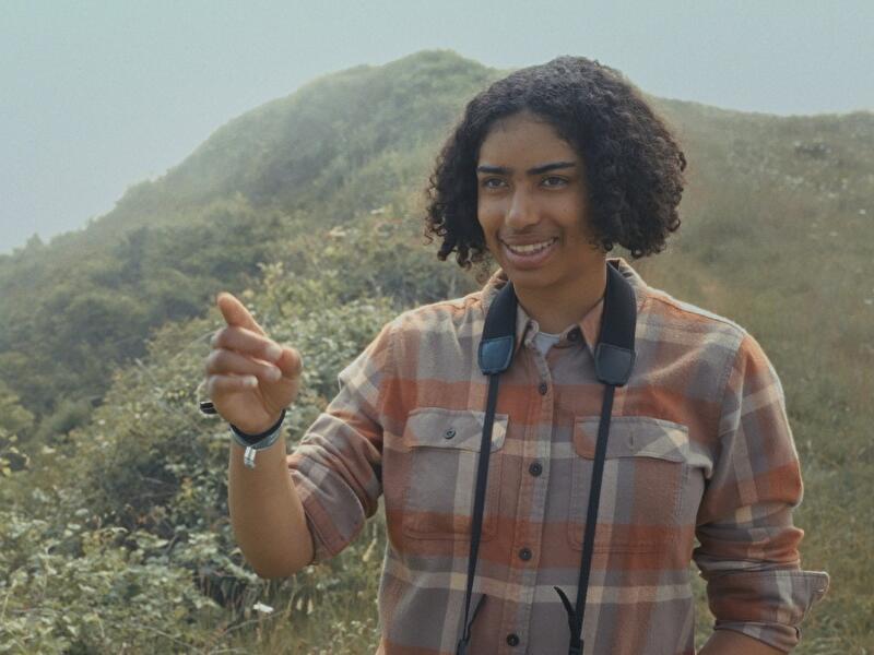 Person mit Fernglas um den Hals steht in hügeliger Landschaft und zeigt auf etwas in der Ferne.
