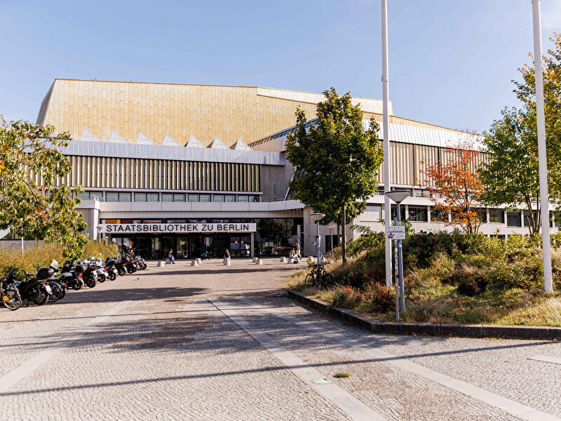 Eingang der Staatsbibliothek zu Berlin mit goldfarbener Fassade, Vorplatz, Bäumen und parkenden Fahrrädern.