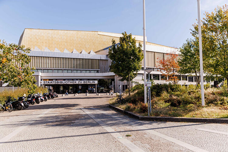 Eingang der Staatsbibliothek zu Berlin mit goldfarbener Fassade, Vorplatz, Bäumen und parkenden Fahrrädern.