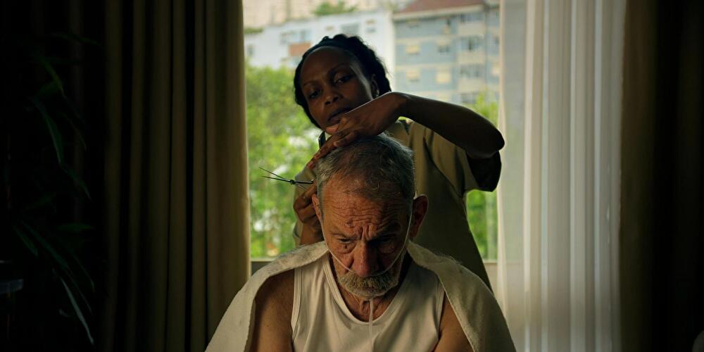 Filmszene aus A Memória do Cheiro das Coisas: Eine Person schneidet einer sitzenden Person mit grauem Haar und Bart die Haare. Die sitzende Person trägt ein weißes ärmelloses Oberteil und hat ein Handtuch über die Schultern gelegt.