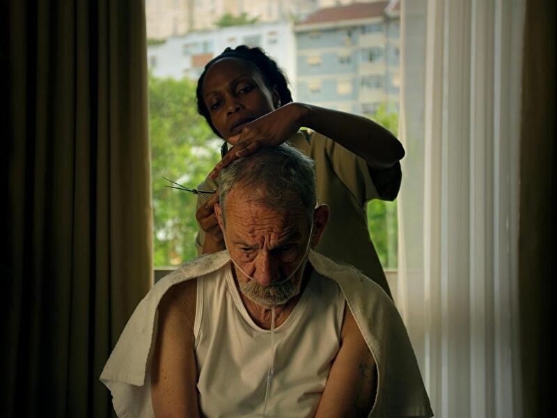 Filmszene aus A Memória do Cheiro das Coisas: Eine Person schneidet einer sitzenden Person mit grauem Haar und Bart die Haare. Die sitzende Person trägt ein weißes ärmelloses Oberteil und hat ein Handtuch über die Schultern gelegt.
