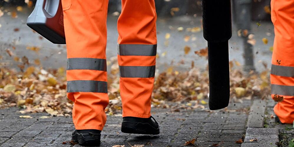 Mitarbeitende der Stadtreinigung beseitigen mit Laubbläsern Herbstlaub auf einem Gehweg.
