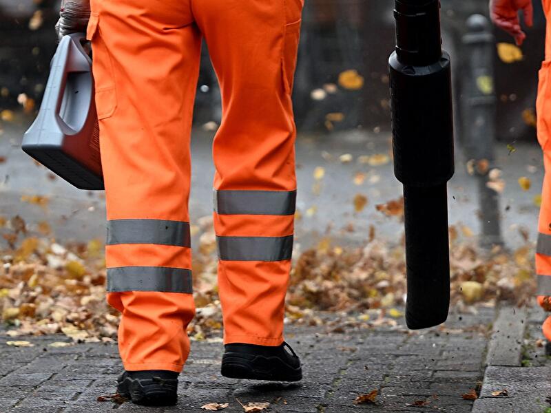 Mitarbeitende der Stadtreinigung beseitigen mit Laubbläsern Herbstlaub auf einem Gehweg.