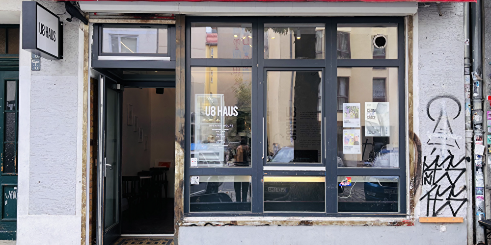 Straßenansicht des U8 Haus, einer Galerie in Berlin. Zu sehen sind die Eingangstür und große Schaufenster unter einer roten Markise.