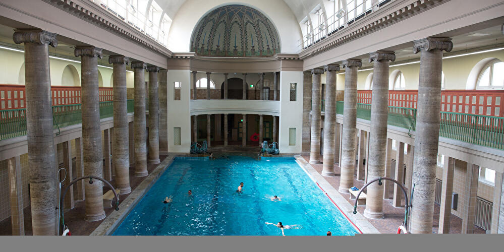 Blick in die historische Schwimmhalle des Stadtbads Neukölln mit großem Becken, hohen Säulenreihen, gewölbter Decke und dekorativer Apsis. Mehrere Badegäste schwimmen im türkisblauen Wasser der lichtdurchfluteten Halle.