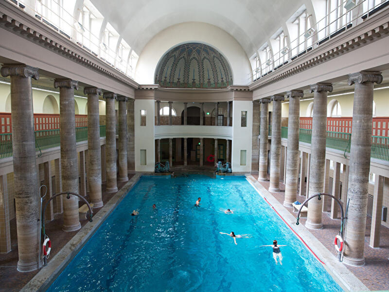 Blick in die historische Schwimmhalle des Stadtbads Neukölln mit großem Becken, hohen Säulenreihen, gewölbter Decke und dekorativer Apsis. Mehrere Badegäste schwimmen im türkisblauen Wasser der lichtdurchfluteten Halle.
