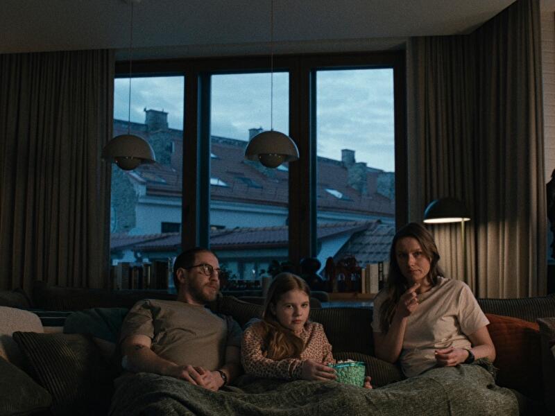 Drei Personen sitzen abends auf einem Sofa vor dem Fernseher, ein Kind hält eine Schüssel Popcorn.