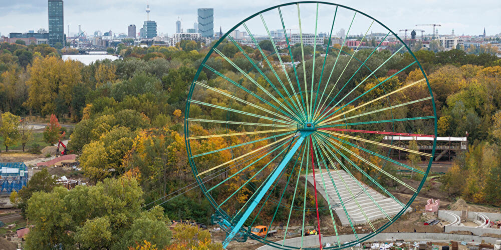 Riesenrad im Spreepark Berlin vor herbstlich gefärbten Bäumen und der Skyline mit Fernsehturm im Hintergrund.