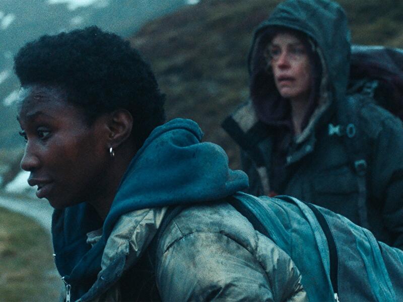 Zwei Personen mit Rucksäcken bewegen sich durch eine raue, bergige Landschaft. Eine Person steht im Vordergrund, die andere folgt in wetterfester Kleidung im Hintergrund.