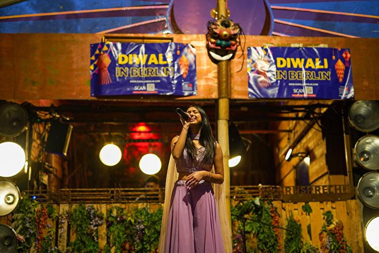 Sängerin tritt auf einer festlich beleuchteten Bühne beim Diwali-Fest in Berlin auf. Im Hintergrund sind Veranstaltungsbanner und farbenreiche Dekorationen zu sehen.