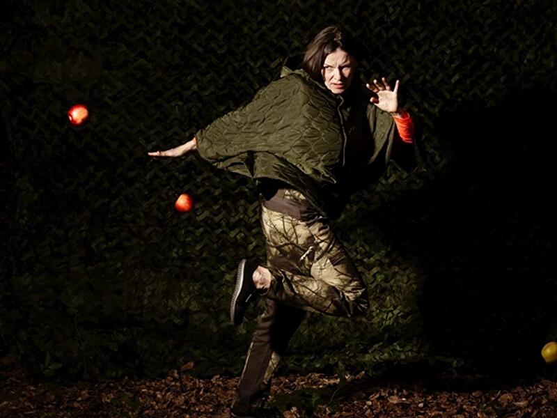 Person in tarnfarbener Kleidung springt vor dunklem Netz-Hintergrund durch eine Performance-Szene. Mehrere leuchtende Bälle schweben in der Luft, dramatisch ausgeleuchtet.