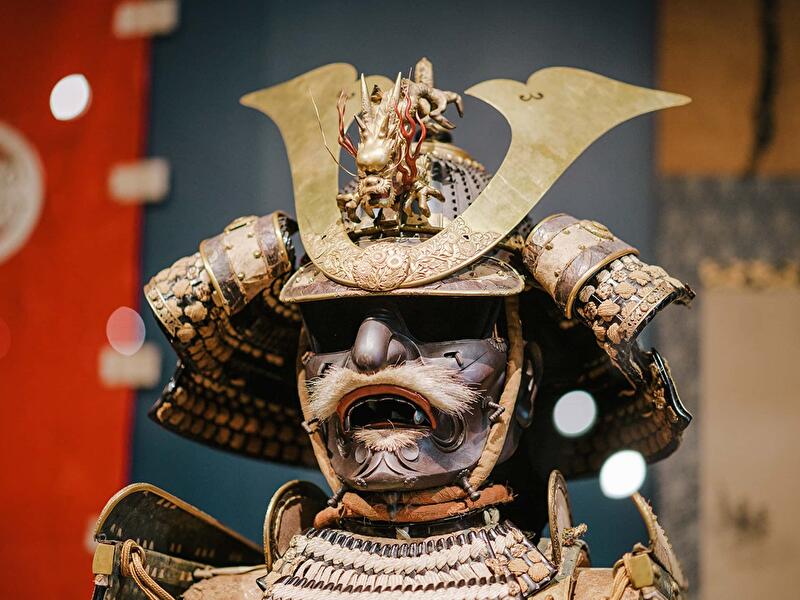 Detailansicht einer historischen Samurai-Rüstung mit verziertem Helm. Auf dem Helm befindet sich ein goldfarbenes Schmuckelement mit Drachenmotiv, darunter eine Gesichtsmaske mit markantem Schnurrbart. Die Rüstung besteht aus kunstvoll gearbeiteten Metall- und Textilsegmenten