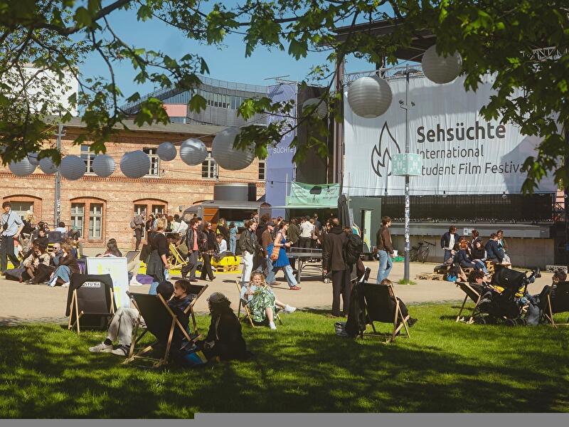 Besucher:innen entspannen auf Liegestühlen und Wiesenflächen vor dem Gelände des Internationalen Studierendenfilmfestivals Sehsüchte. Im Hintergrund sind Veranstaltungsbereiche, historische Backsteinbauten und ein großes Festivalbanner zu sehen.