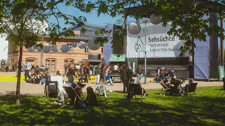 Besucher:innen entspannen auf Liegestühlen und Wiesenflächen vor dem Gelände des Internationalen Studierendenfilmfestivals Sehsüchte. Im Hintergrund sind Veranstaltungsbereiche, historische Backsteinbauten und ein großes Festivalbanner zu sehen.