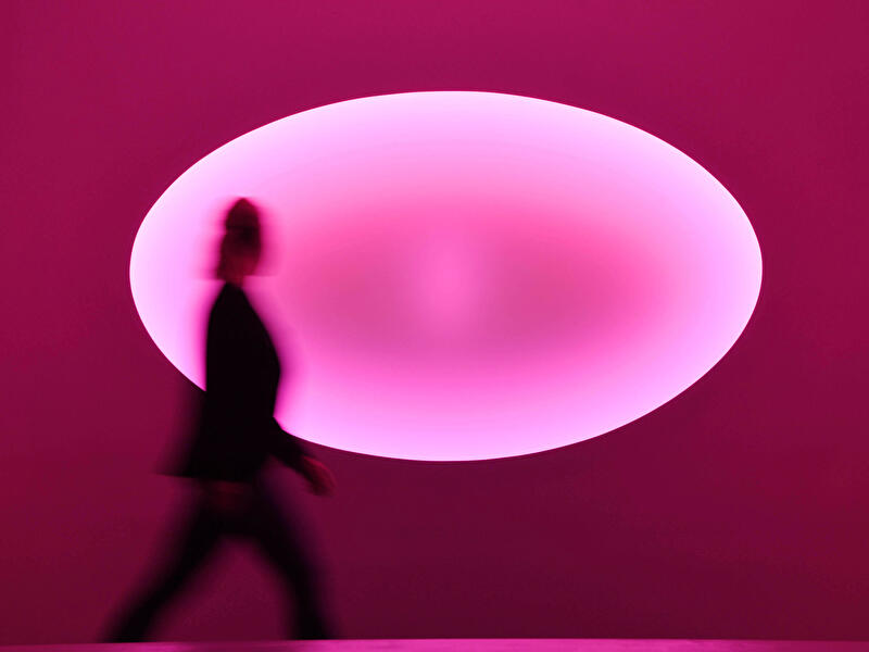Silhouette einer gehenden Person vor einer großen ovalen, pink leuchtenden Lichtinstallation des Künstlers James Turrell an der Wand
