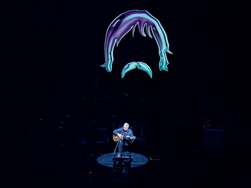 Eine Person sitzt allein auf einer dunklen Bühne im Scheinwerferlicht und spielt Gitarre. Über der Bühne ist die stilisierte Zeichnung eines Kopfes mit Haaren und Schnurrbart projiziert.