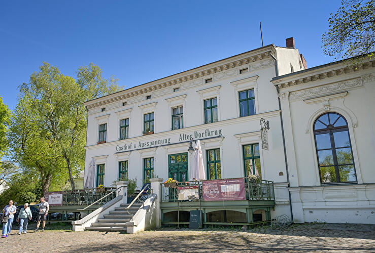 Gasthof Alter Dorfkrug in Berlin-Lübars mit Terrasse, Freitreppe und alten Bäumen unter blauem Himmel.
