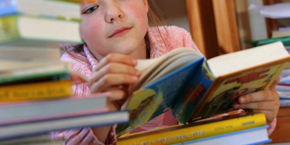 Mädchen blättert in Buch umgeben von Bücherstapel