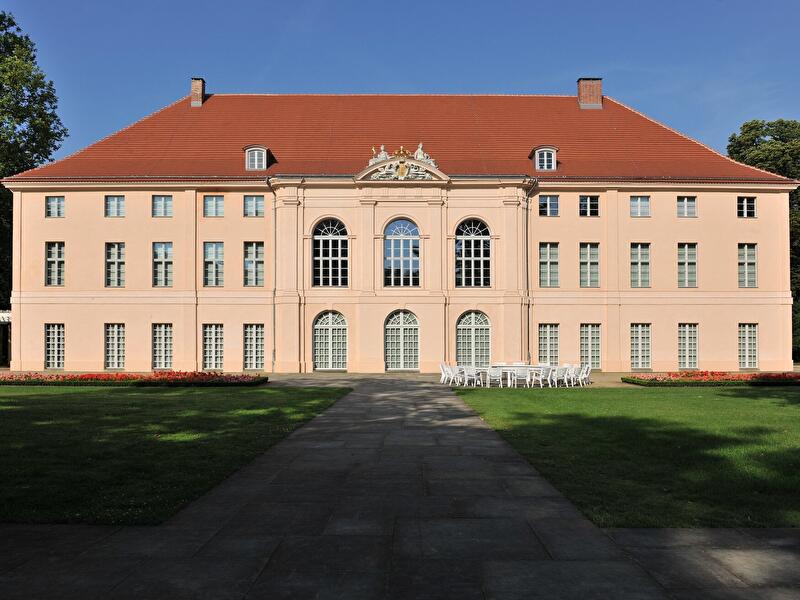 Frontalansicht des barocken Schlosses Schönhausen in Berlin. Das hellrosa Gebäude mit rotem Dach steht hinter einer gepflegten Rasenfläche. 