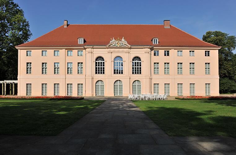 Frontalansicht des barocken Schlosses Schönhausen in Berlin. Das hellrosa Gebäude mit rotem Dach steht hinter einer gepflegten Rasenfläche. 