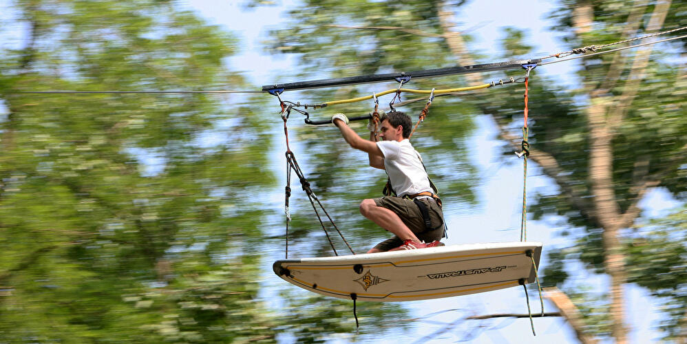 Person auf Surfbrett gleitet an Seilbahn durch einen Kletterwald zwischen Bäumen.