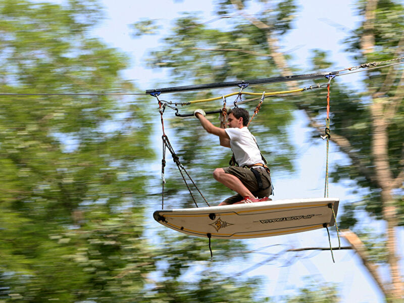 Person auf Surfbrett gleitet an Seilbahn durch einen Kletterwald zwischen Bäumen.