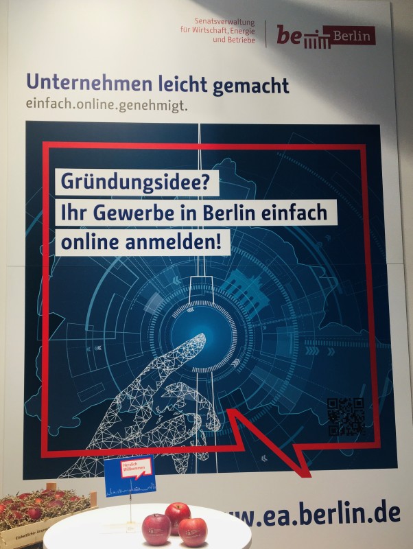 Plakat zur Gewerbeanmeldung beim Stand des EA Berlin auf der deGUT 2019, davor bedruckte Äpfel