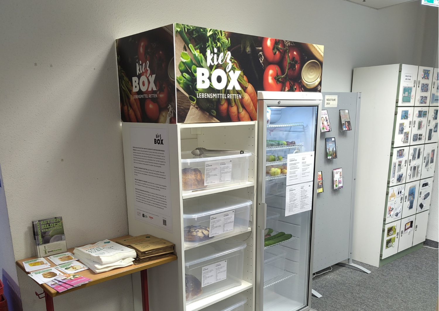 In einem Raum steht ein Retterkühlschrank sog. Kiezbox daneben ein Tisch mit Infomaterial