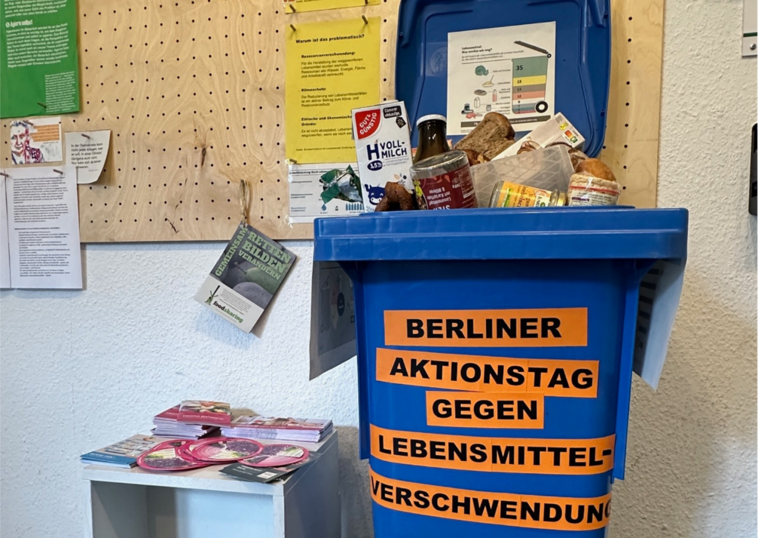 Mülltonne mit der Aufschrift Berliner Aktionstag gegen Lebensmittelverschwendung steht vor einer großen Pinnwand
