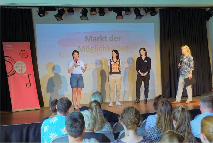 Blick aus einem sitzenden Publikum auf eine Bühne, auf der vier Personen stehen