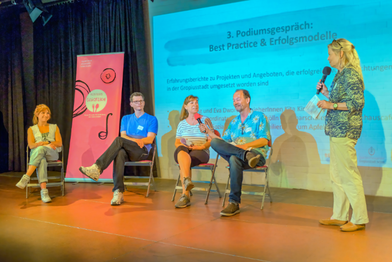 fünf Personen nehmen auf einer Bühne an einem Podiumsgespräch teil, vier Personen sitzen und eine Frau moderiert im Stehen