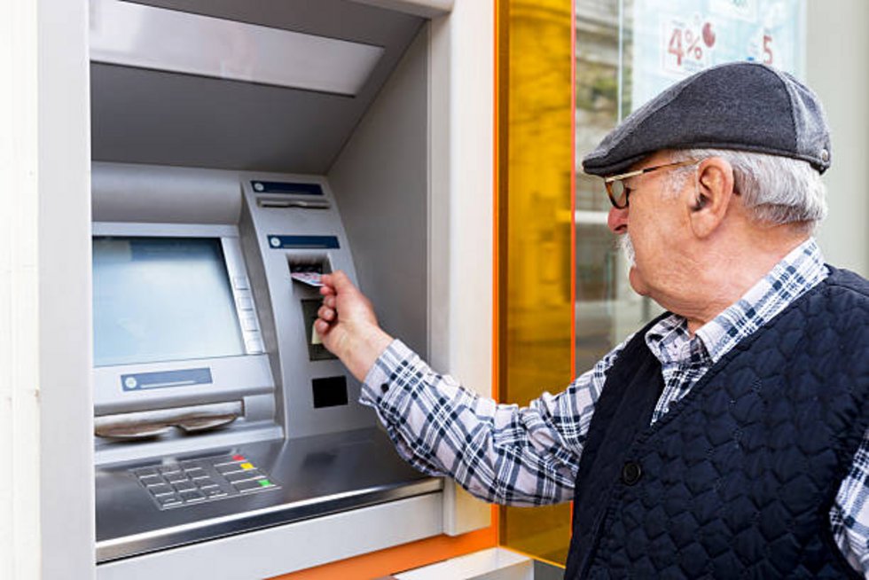 Grauhaariger Mann im Karohemd und Schiebermütze steckt Karte in einen Geldautomaten.