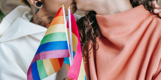 Zwei Frauen küssen sich und halten beide die Regenbogenflagge vor sich hoch.