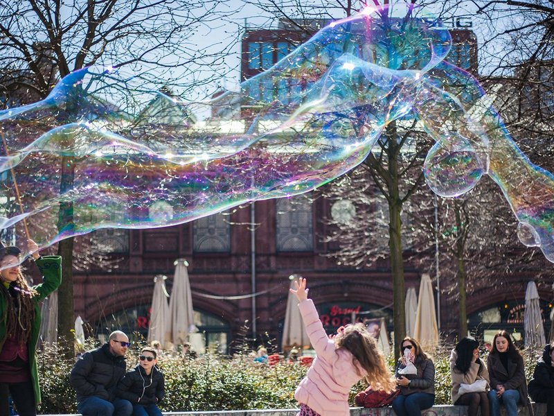 Menschen halten sich bei Sonnenschein auf einem städtischen Platz auf. Eine Frau macht riesige Seifenblasen, ein Mädchen tanzt darunter hindurch