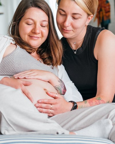 Zwei Frauen auf einer gelben Couch, die Frau links hat einen Babybauch, den beide mit den Händen berühren
