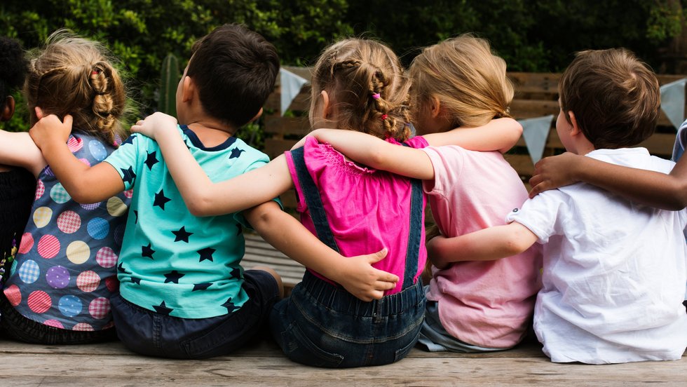 Eine Gruppe Kitakinder umarmt sich auf einem Holzbalken sitzend, von hinten zu sehen.