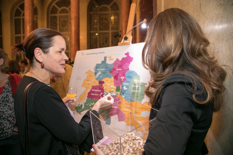 Eine Frau mit einem Behälter voller Pinnadeln im Gespräch mit einer weiteren Frau vor der Gender-Map