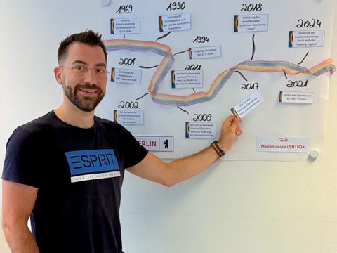 Das Foto zeigt einen Mitarbeiter des Bezirksamts Friedrichshain-Kreuzberg, der in der Queeren Aktionswoche an einem Quiz zu den Meilensteinen der LSBTIQ+ teilnimmt und vor einem Plakat steht.