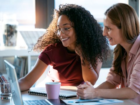 Zwei lächelnde Frauen schauen zusammen auf einen Laptop in einer Büroumgebung