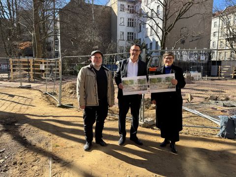 Drei Personen stehen auf einem Baustellengelände, zwei halten Pläne mit Landschaftsbildern