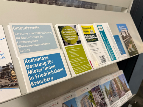 Mehrere Flyer liegen nebeneinander auf einem weißen Regal in einem Flur. Im Vordergrund befindet sich ein Flyer mit der Überschrift 'Kostenlose Beratung für Mieter*innen in Friedrichshain-Kreuzberg'. Daneben sind weitere Informationsflyer zu Themen wie Ombudsstelle, Mietrecht und Wohnungsunternehmen in Berlin ausgelegt. Die Flyer sind farbig gestaltet, überwiegend in Blau-, Grün- und Gelbtönen. Im Hintergrund sind weitere Broschüren sichtbar, die thematisch an die Dokumentation von Bauanträgen und Erhaltungsgebieten im Bezirksamt Friedrichshain-Kreuzberg anschließen.