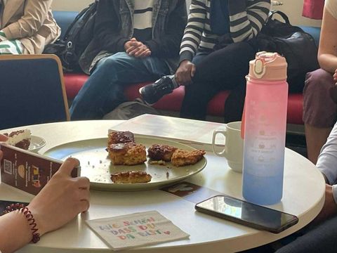 Menschen sitzen zusammen an einem Tisch. Auf dem Tisch stehen Kuchenstücke, Tassen und ein Infozettel vom Sprach-Café.