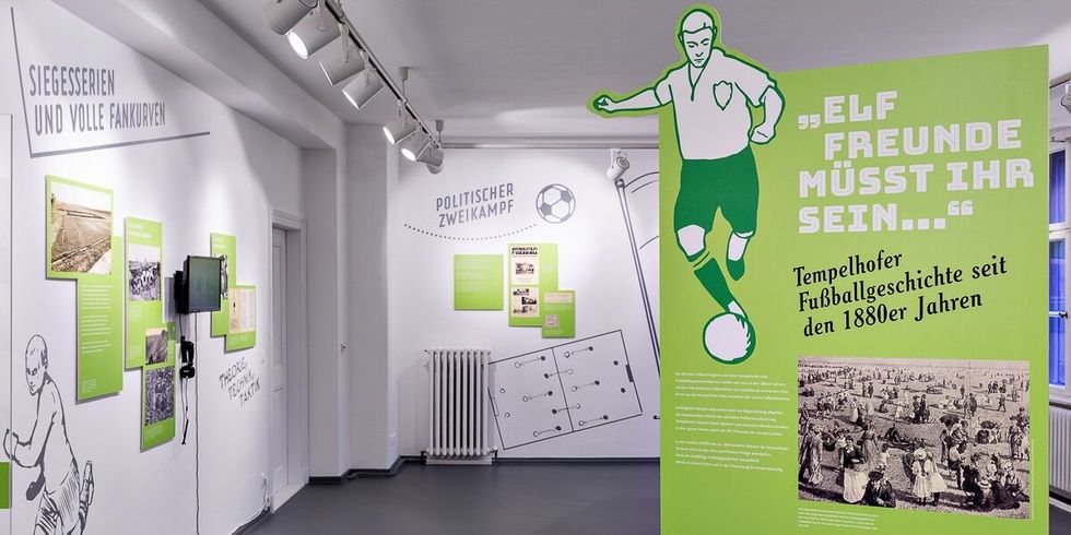 In einem weißen Raum sind Infotafeln und Fußballbilder an den Wänden. Im Raum steht eine Infotafel mit einem Fußballer und dem Textzug "Elf Freunde müsst ihr sein..." Tempelhofer Fußballgeschichte seit den 1880er Jahren.