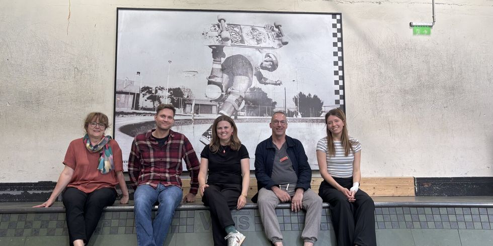 Fünf Personen sitzen nebeneinander auf dem Rand einer Skateboard-Rampe in einer Halle. Im Hintergrund hängt ein großes Schwarz-Weiß-Foto, das einen Skateboarder bei einem Trick zeigt. Die Gruppe wirkt entspannt und lächelt in die Kamera. Die Wände sind schlicht, und über dem Foto ist ein Notausgangsschild zu sehen.