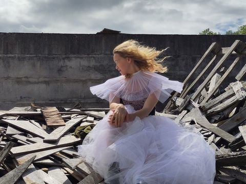 Ein junges Mädchen sitzt in einem Rüschenkleid nachdenklich auf einem Haufen Bauholz vor einer Mauer im Freien