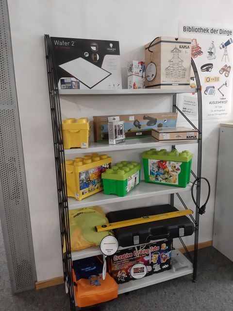 Regal "Bibliothek der Dinge" gefüllt mit verschiedenen Dingen, wie Kinderteleskop, Lego-Box, Wasserwage und Metalldetektor