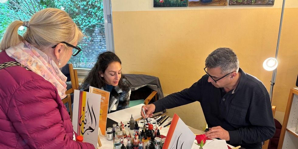 Ein Mann malt arabische Kaligraphie zwei Frauen beobachten den Künstler am Tisch